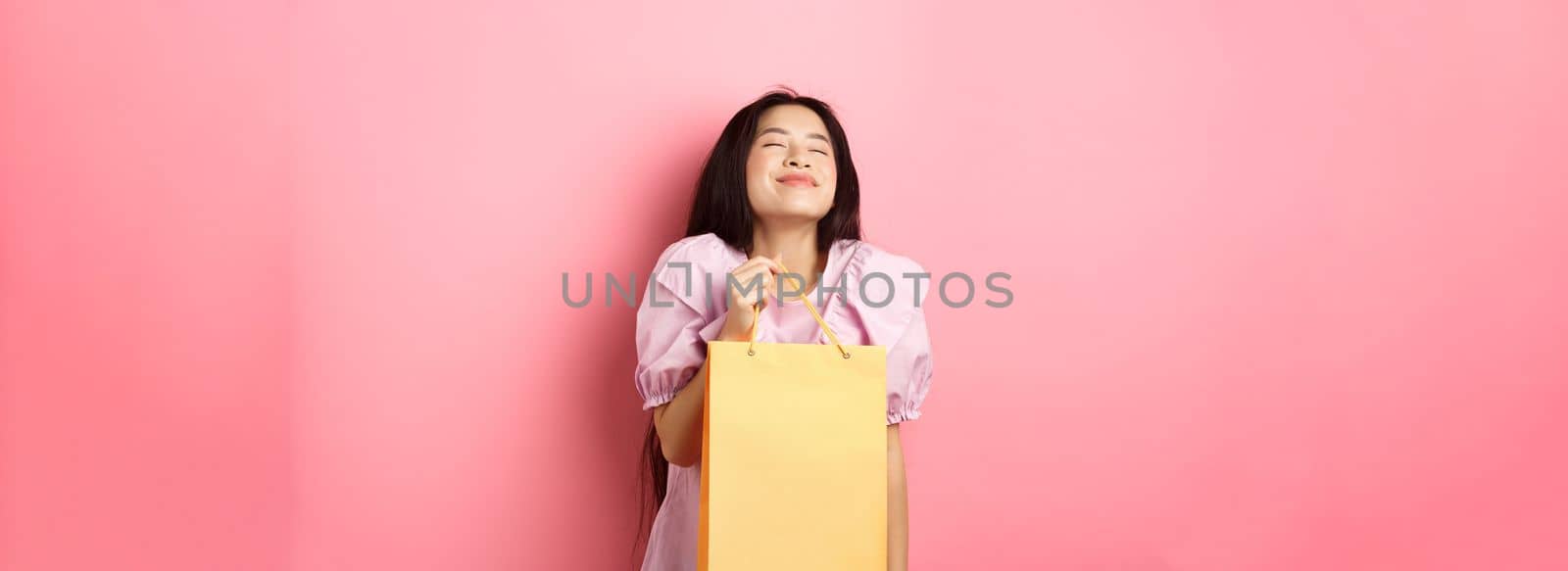 Beautiful happy asian woman holding shopping bag, smiling with eyes closed, standing in dress against pink background by Benzoix