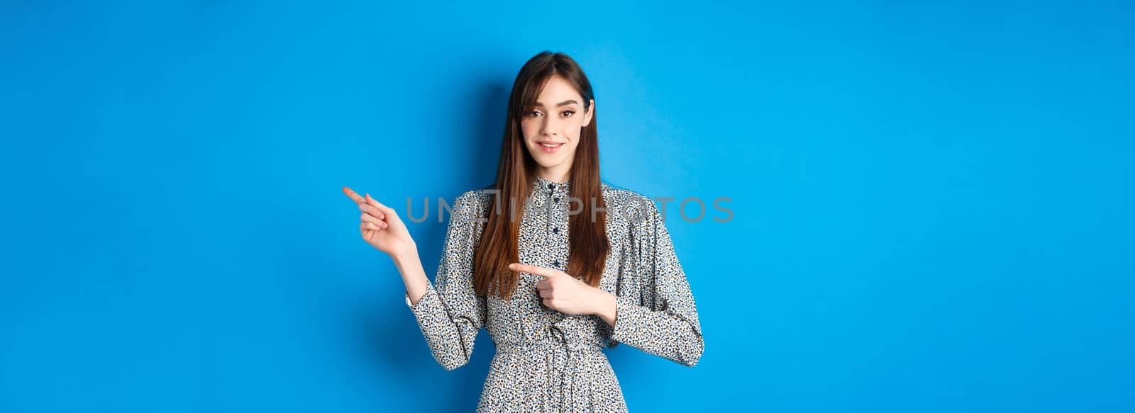 Young attractive woman in dress, with long natural hair, pointing fingers right at logo, smiling and inviting check out promotion, standing on blue background by Benzoix