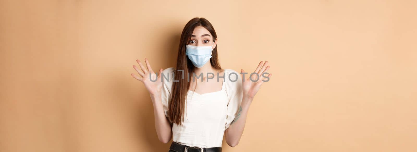 Covid-19 and lifestyle concept. Surprised woman in medical mask scream excited, raising hands up and shouting big news, standing on beige background.