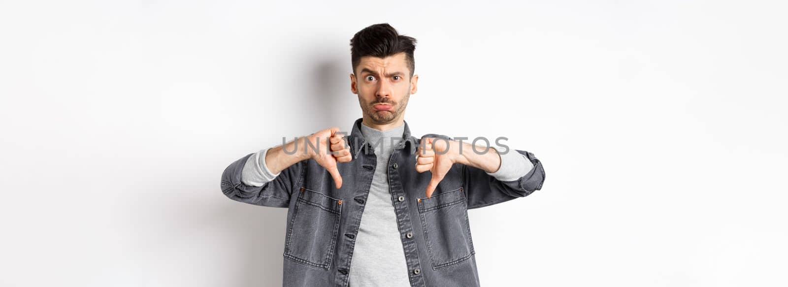 Disappointed funny man frowning and looking upset, showing thumbs down displeased, standing on white background.