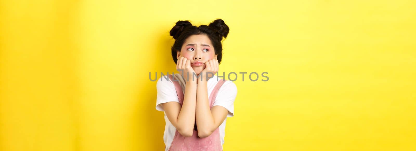 Sad moody asian girl, lean face on hands, looking left with sulking expression, standing in summer clothes on yellow background by Benzoix