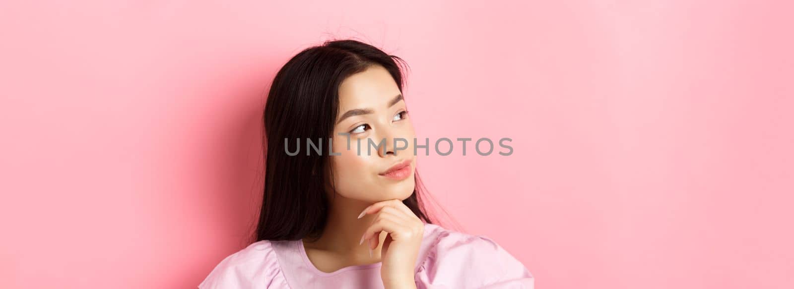 Close-up portrait of romantic asian girl looking aside at logo, thinking or daydreaming, standing against pink background.