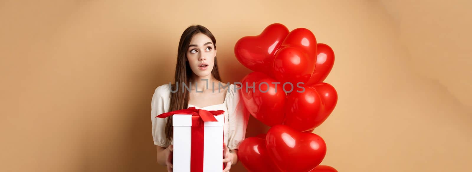 Valentines day. Pensive cute girl guessing who made her gift, holding present and looking curious at upper left corner, standing near romantic heart balloons, beige background by Benzoix