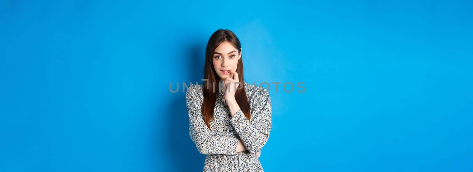 Sensual woman in dress biting finger and looking pensive at camera, standing dreamy against blue background.