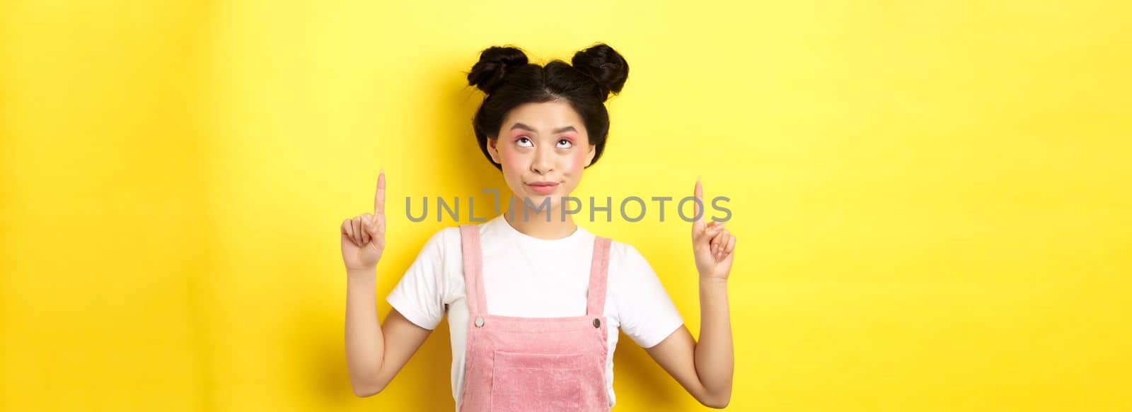 Glamour asian girl with beauty makeup, pointing fingers up, showing top advertisement, standing on yellow background.