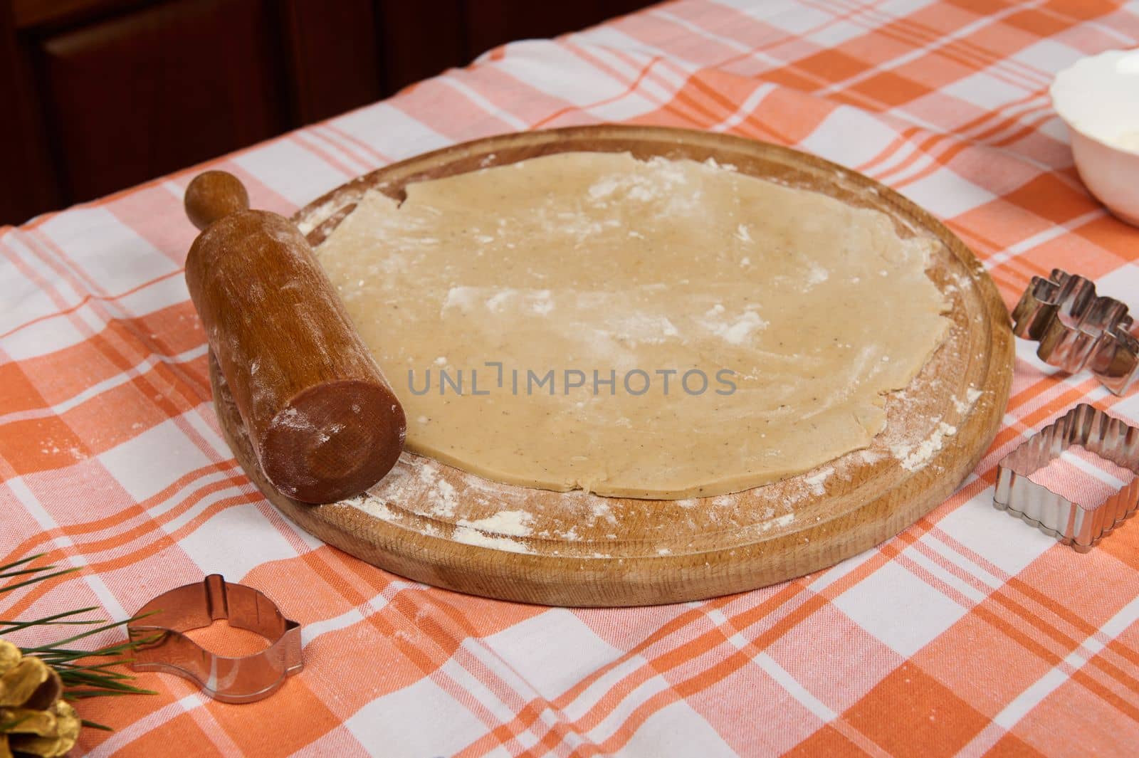 Still life with floured rolled out gingerbread dough and rolling pin on a wooden board. Cookies cutters on the red plaid tablecloth