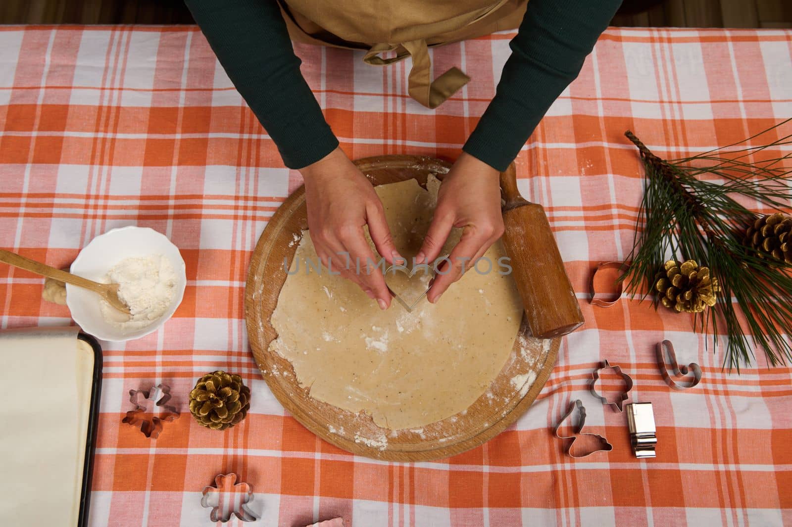 View from above a housewife, female chef confectioner holding a heart shaped cookie cutter in her hands, carving molds on the gingerbread dough, preparing traditional Christmas cookies. Baking concept