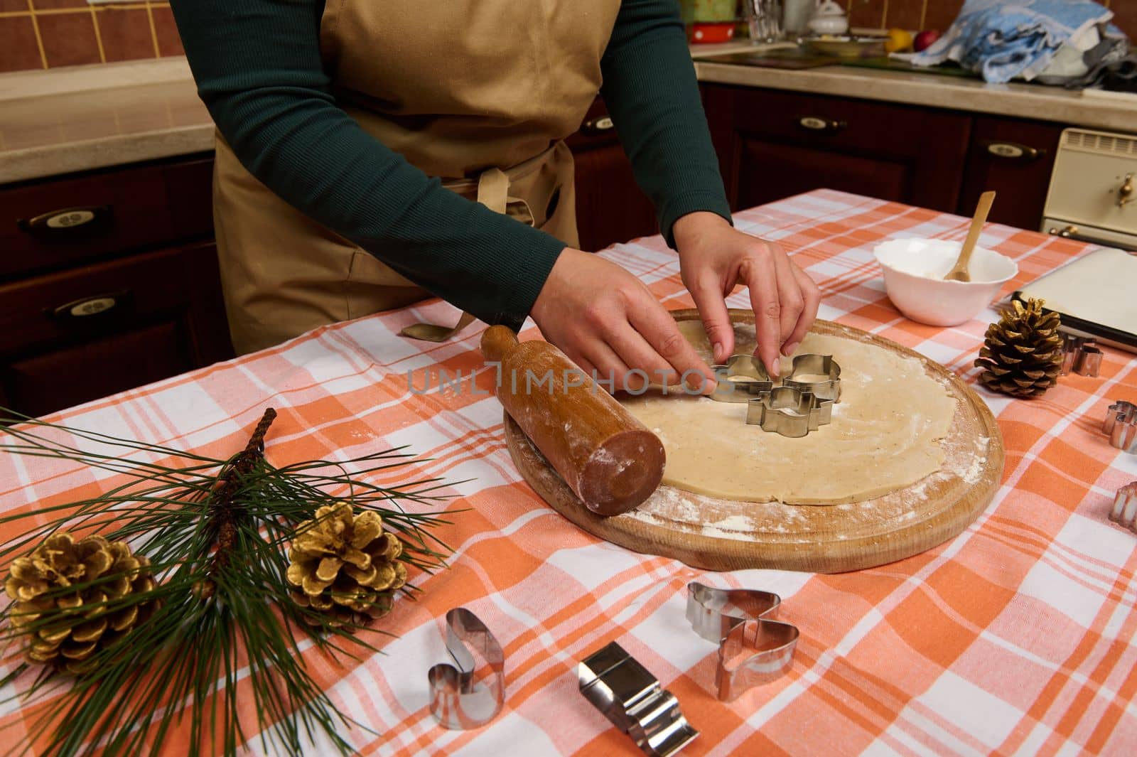 Close-up female confectioner in beige chef's apron, metal putting cookies cutters on a rolled out gingerbread dough, preparing delicious homemade pastries for Christmas holidays in the kitchen at home