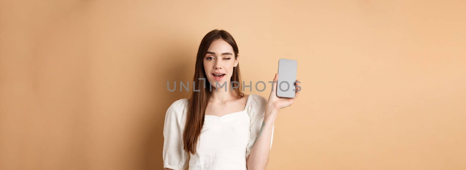 Cheerful stylish girl showing smartphone app, demonstrate empty phone screen and winking, standing on beige background.