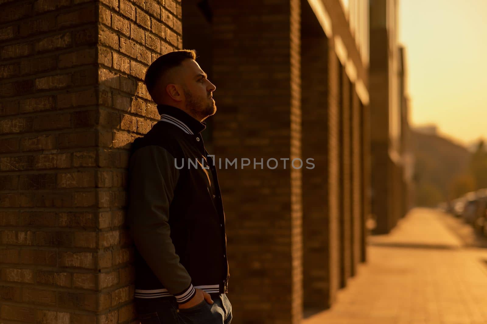 Caucasian bearded man in a bomber jacket leaned against a brick wall