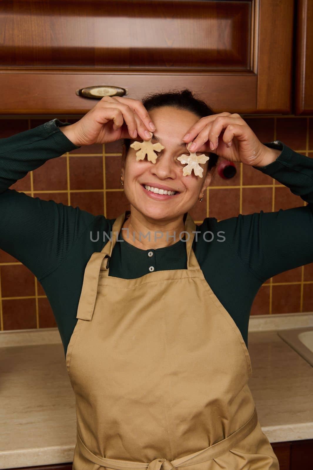 Cheerful housewife in green shirt and beige apron, holding two snowflake shaped gingerbread dough near her eyes and smiling a beautiful toothy smile, while baking christmas cookies in the home kitchen