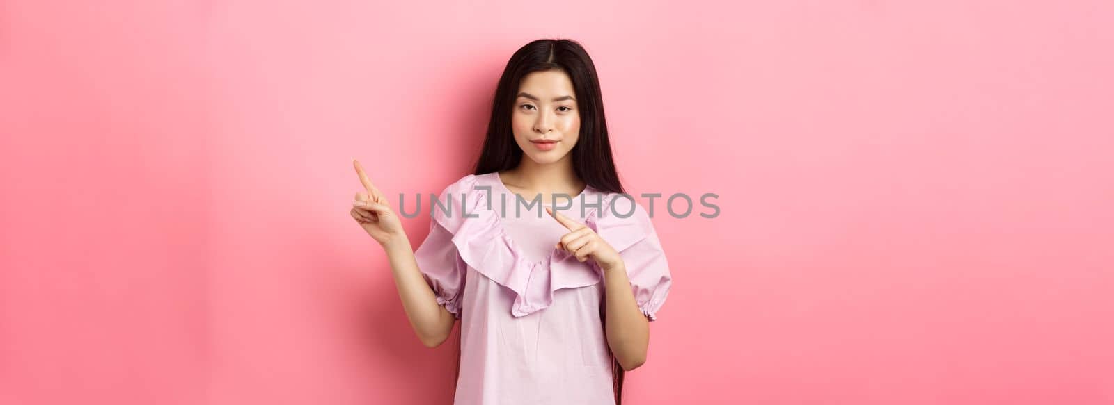 Cheeky asian girl showing interesting advertisement, pointing fingers right at logo and smiling cunning, hinting on good deal, pink background by Benzoix
