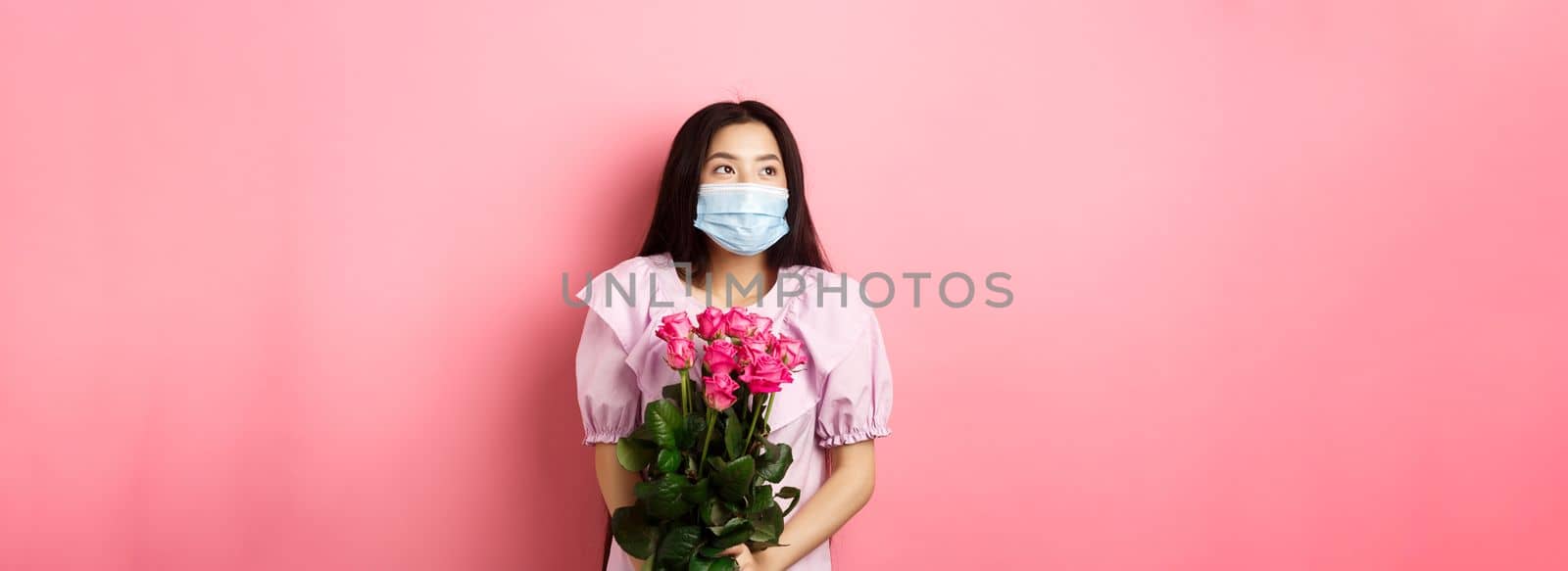 Romantic asian girl in medical mask looking aside at empty space with dreamy eyes, holding bouquet of roses for Valentines day, having date with lover during pandemic by Benzoix