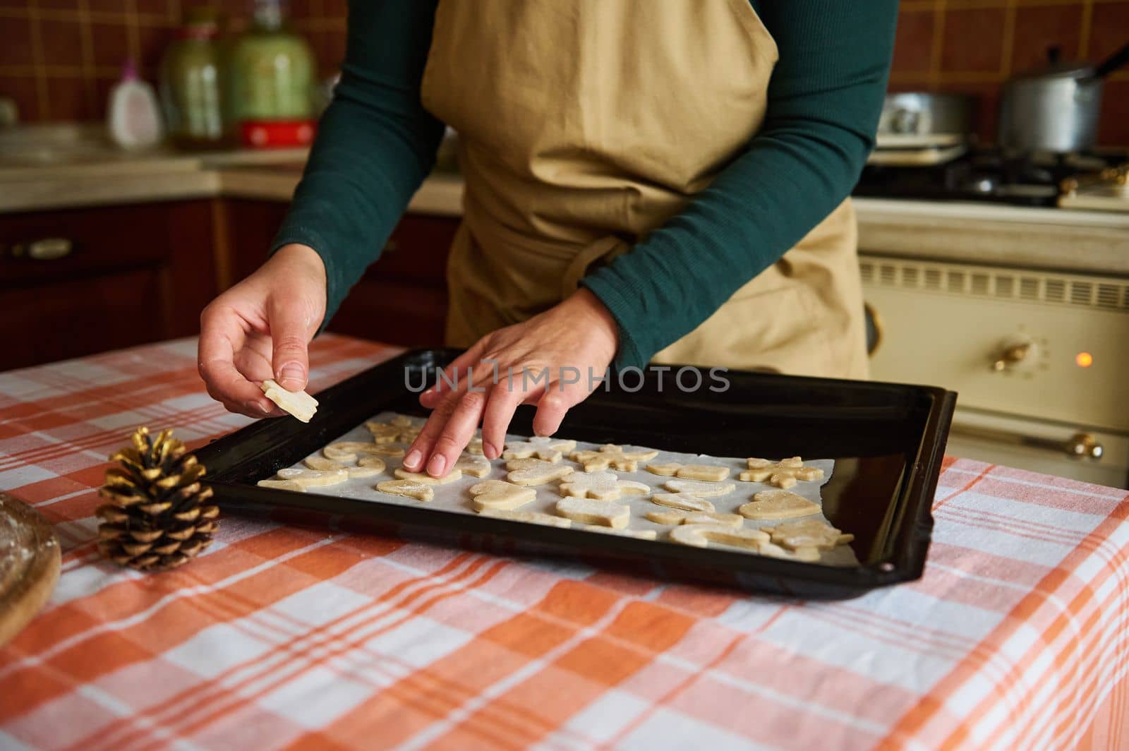 A woman housewife in beige chef's apron, putting cookies cut out of gingerbread dough on the baking sheet, preparing Christmas pastries in the home kitchen with wooden vintage design. Baking concept