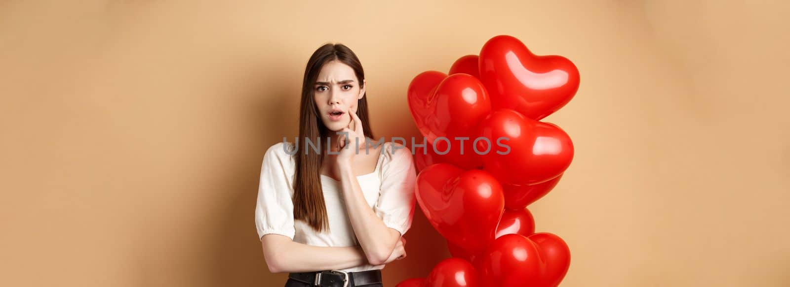 Valentines day and love concept. Confused and shocked girl staring displeased at camera, standing near romantic red hearts balloons on beige background.