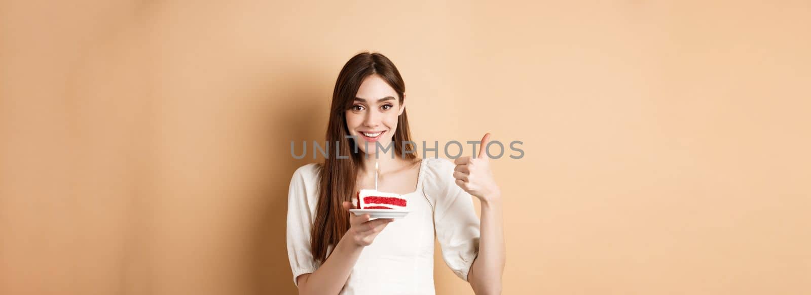 Beautiful birthday girl making wish on cake, showing thumb up and smiling happy, enjoying b-day party, standing on beige background.