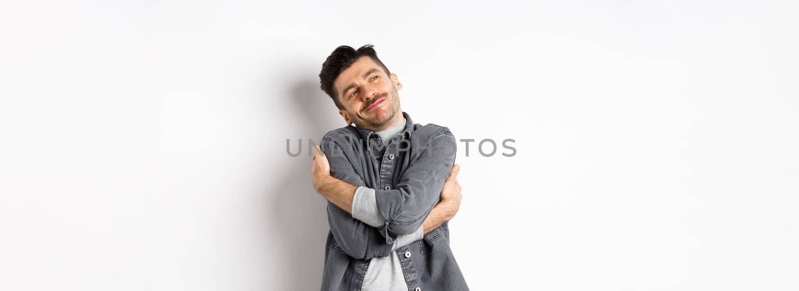 Happy romantic guy hugging himself with closed eyes and smile, loving own body, daydreaming about lover, standing on white background.