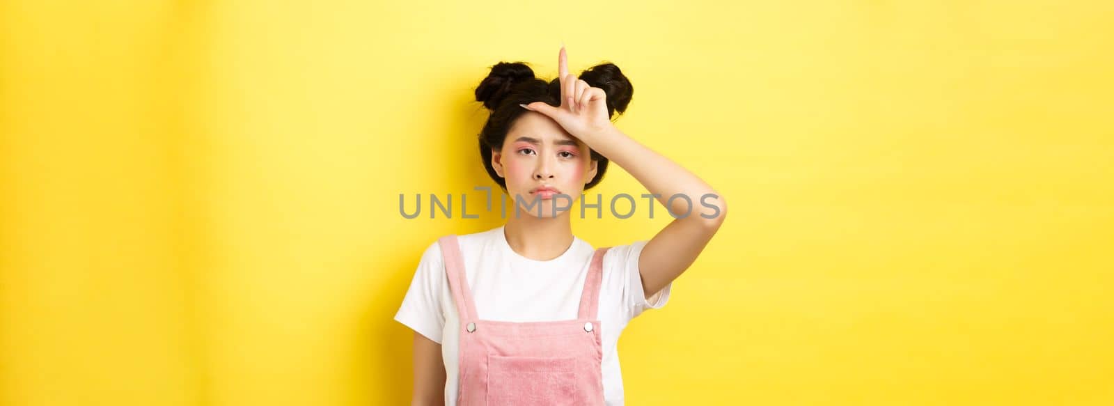 Sad girl showing loser sign on forehead and sulking upset, feeling disappointed in herself, standing on yellow background by Benzoix