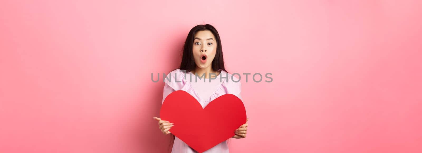 Valentines day concept. Surprised asian teen girl say wow, showing big red heart card, receive gift from secret admirer or lover, standing in romantic dress on pink background by Benzoix