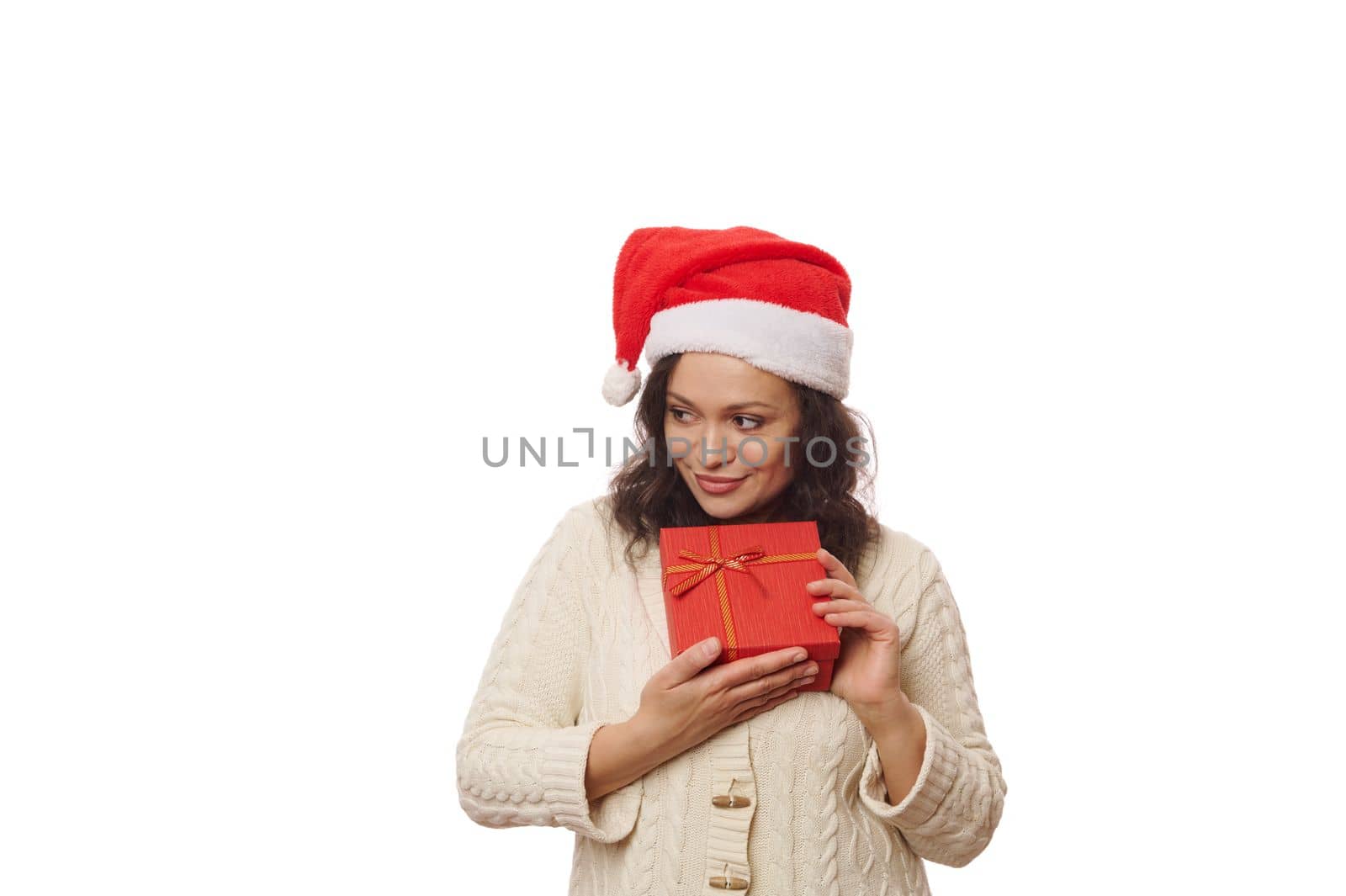 Beautiful African American woman wearing Santa hat and beige sweater, holding Christmas present over white background. Boxing Day. Time to open gift boxes. New Year's preparations. Christmastime