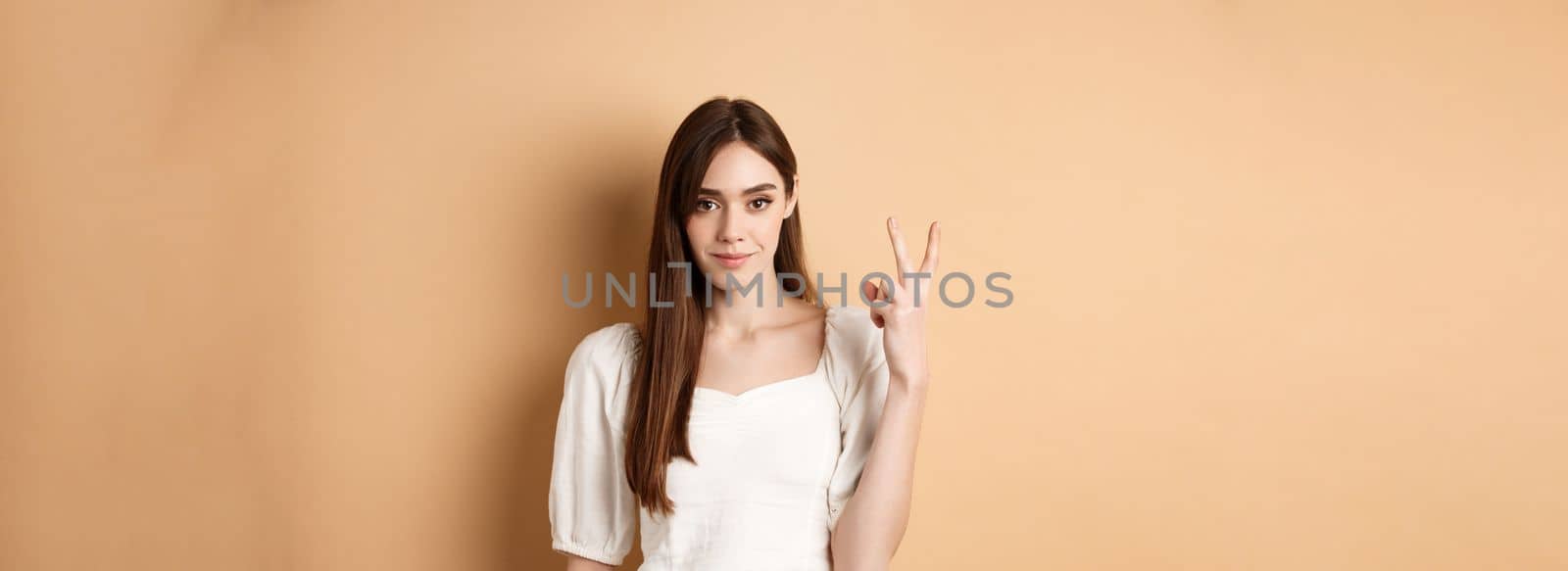 Attractive young woman show fingers number two, smiling and looking confident, standing on beige background.
