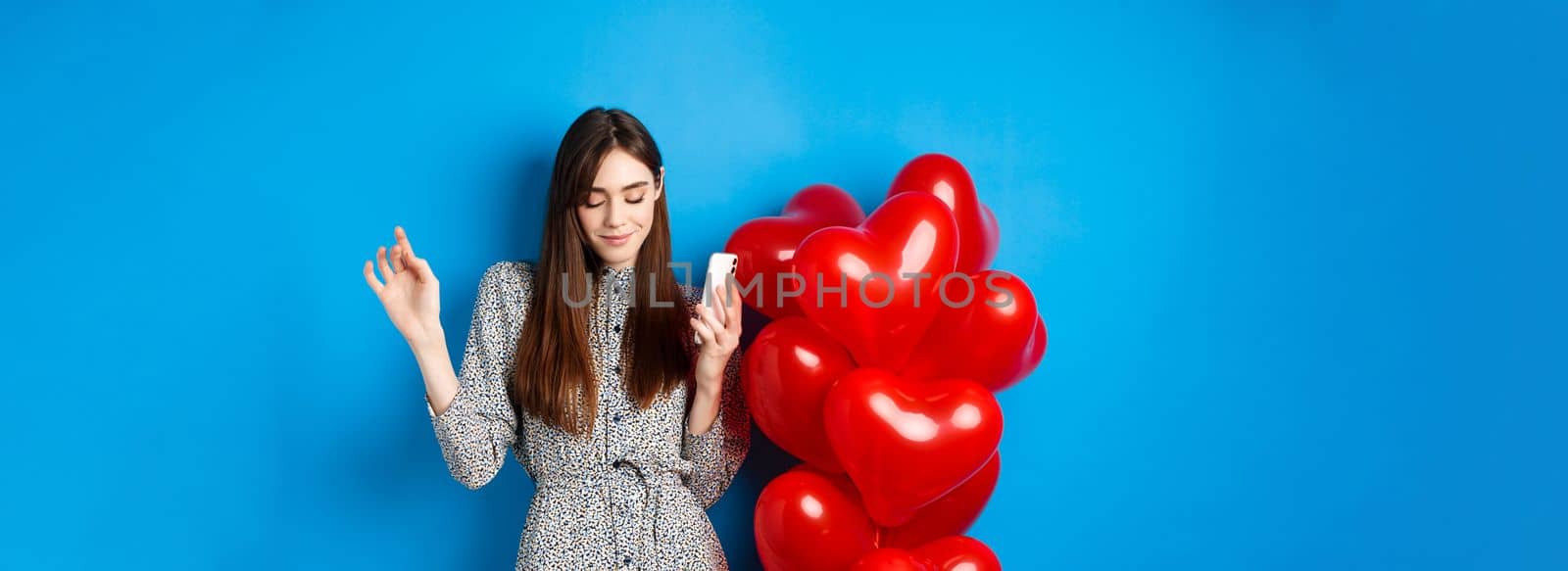 Portrait of attractive young woman holding smartphone and dancing near Valentines red balloons, standing on blue background by Benzoix