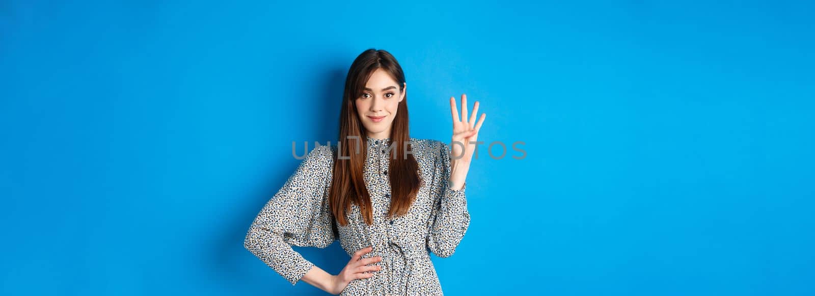 Young beautiful girl in dress with natural long hair, showing number four with fingers and smiling, standing against blue background.