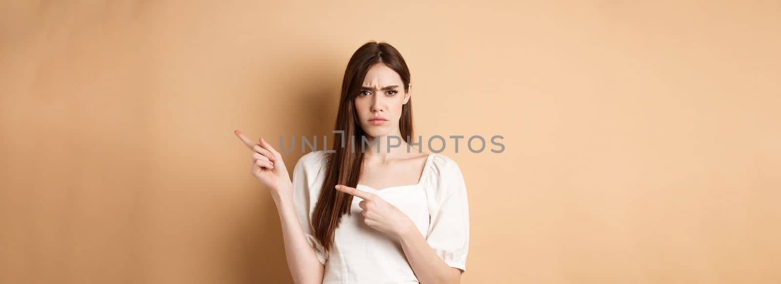 Angry young woman frowning, pointing fingers right and complaining, disappointed with logo, standing upset on beige background by Benzoix