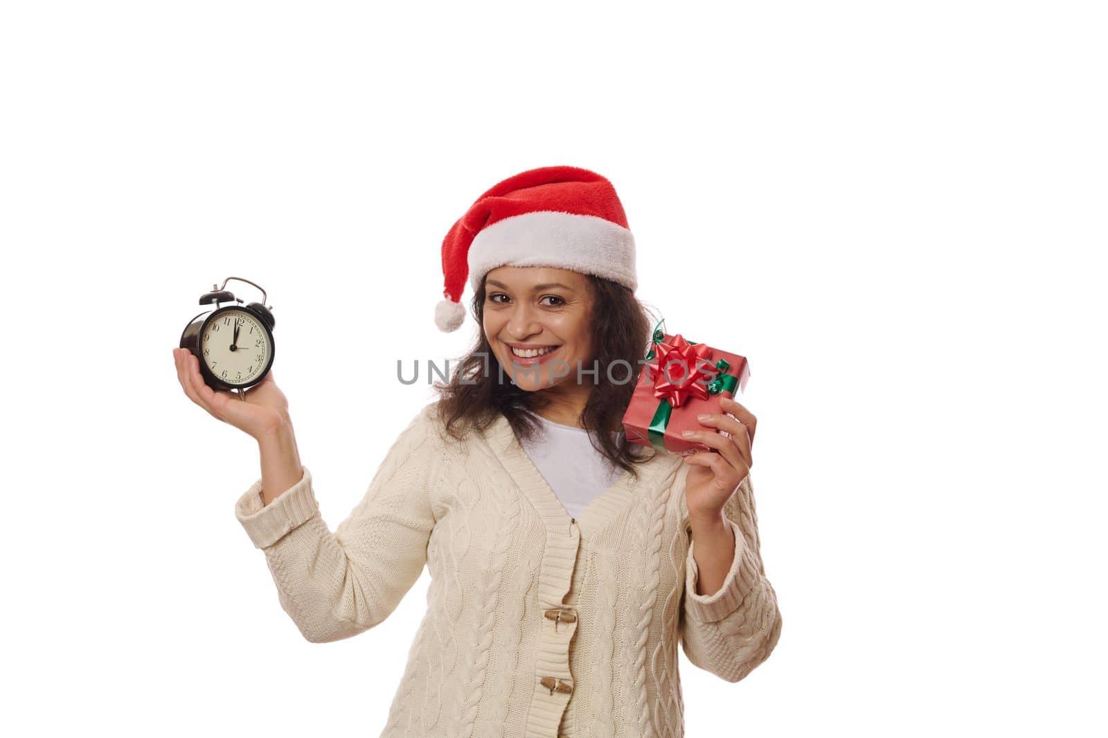 Delightful woman wearing Santa hat, holding a happy Xmas gift box with surprise, and black retro alarm clock showing five minutes to midnight on dial, smiles looking at camera, over white background