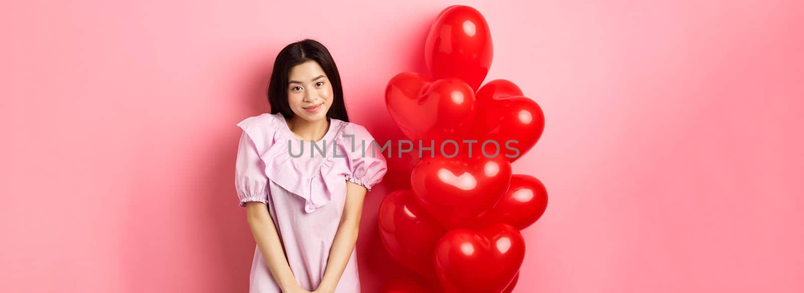 Cute asian girl in dress looking shy and smiling, standing modest near valentines day balloons, blushing on romantic date, looking at camera, pink background.