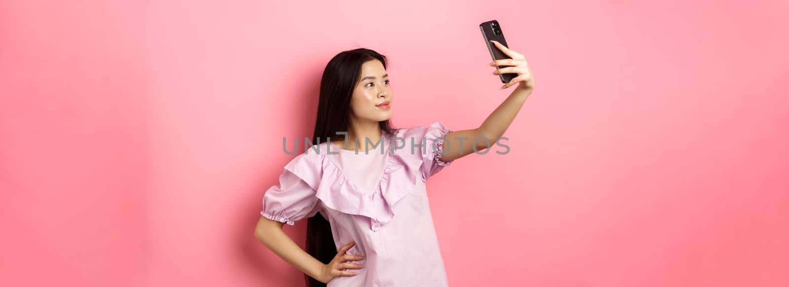 Stylish asian girl taking selfie and smiling, posing for social media photo, standing in dress against pink background.