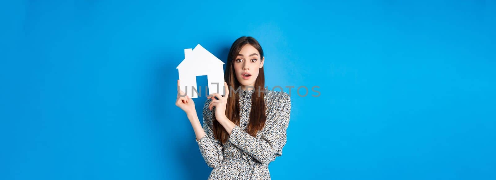 Real estate, realtors and insurance concept. Excited woman gasping and holding paper house cutout, see good deal, standing on blue background.