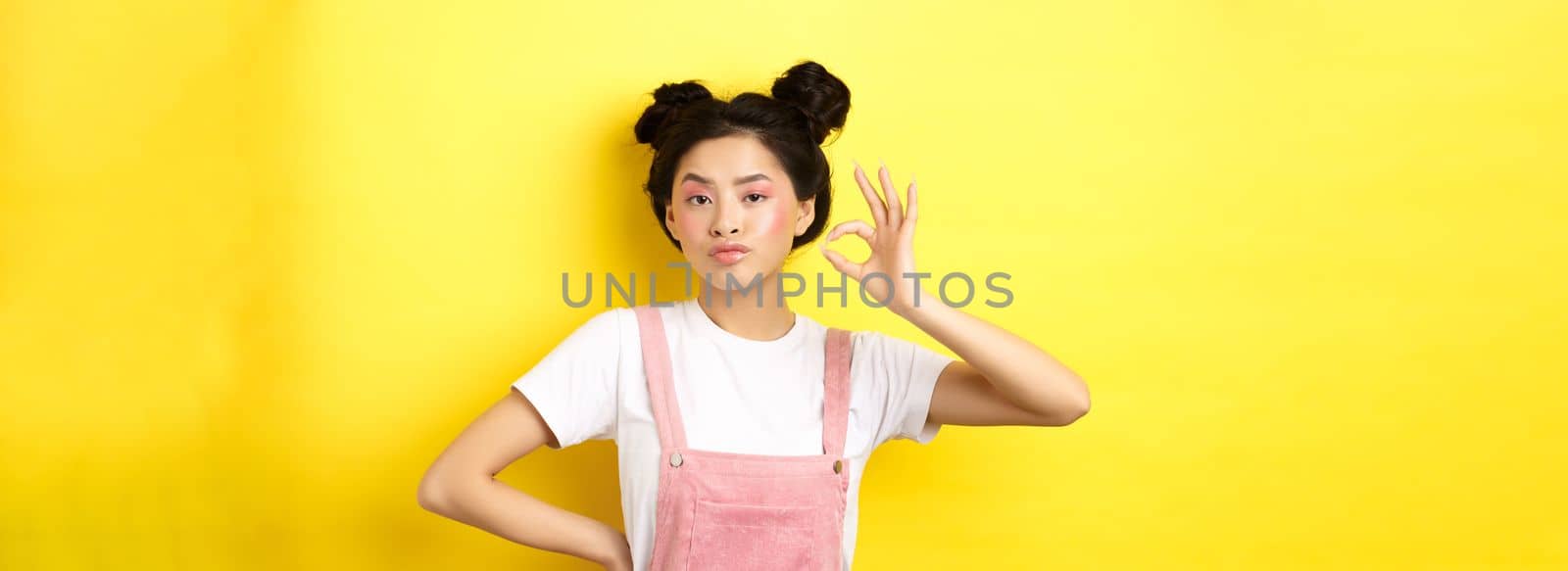 Sassy teenage asian girl with bright pink makeup, showing okay sign and nod in approval, approve product with serious face, not bad gesture, standing against yellow background.