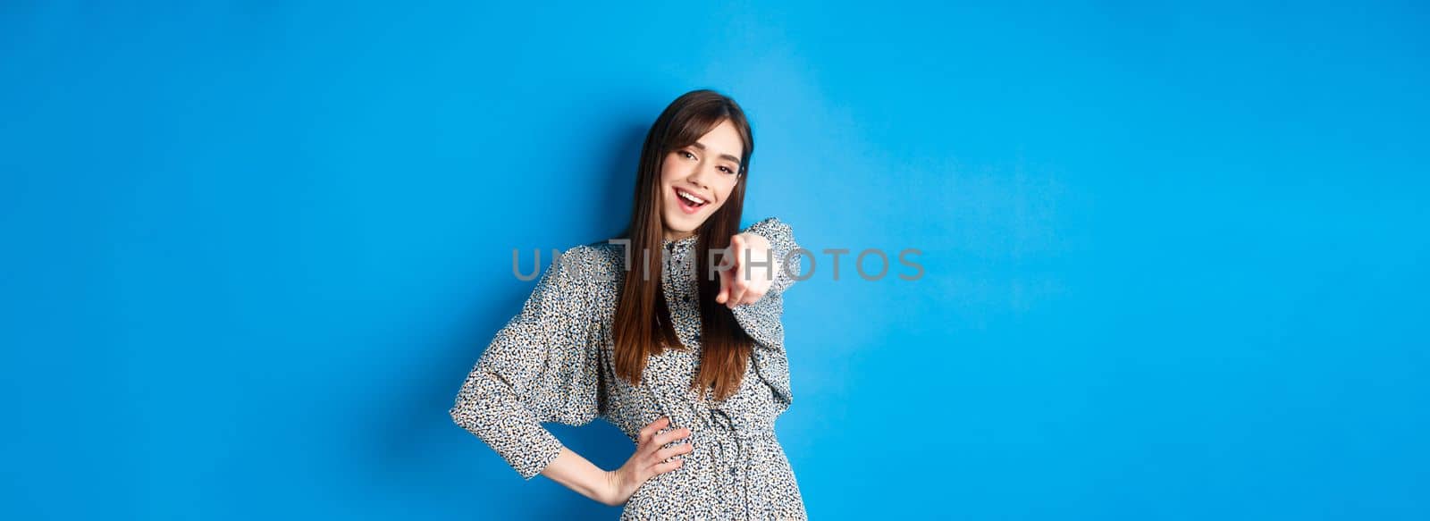 Motivated and excited young woman pointing finger at camera and smiling, congratulating you, praising or inviting, standing happy on blue background.