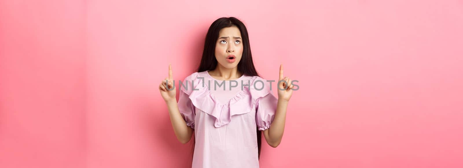 Concerned and sad asian girl frowning, looking and pointing fingers up at bad news, standing against pink background.