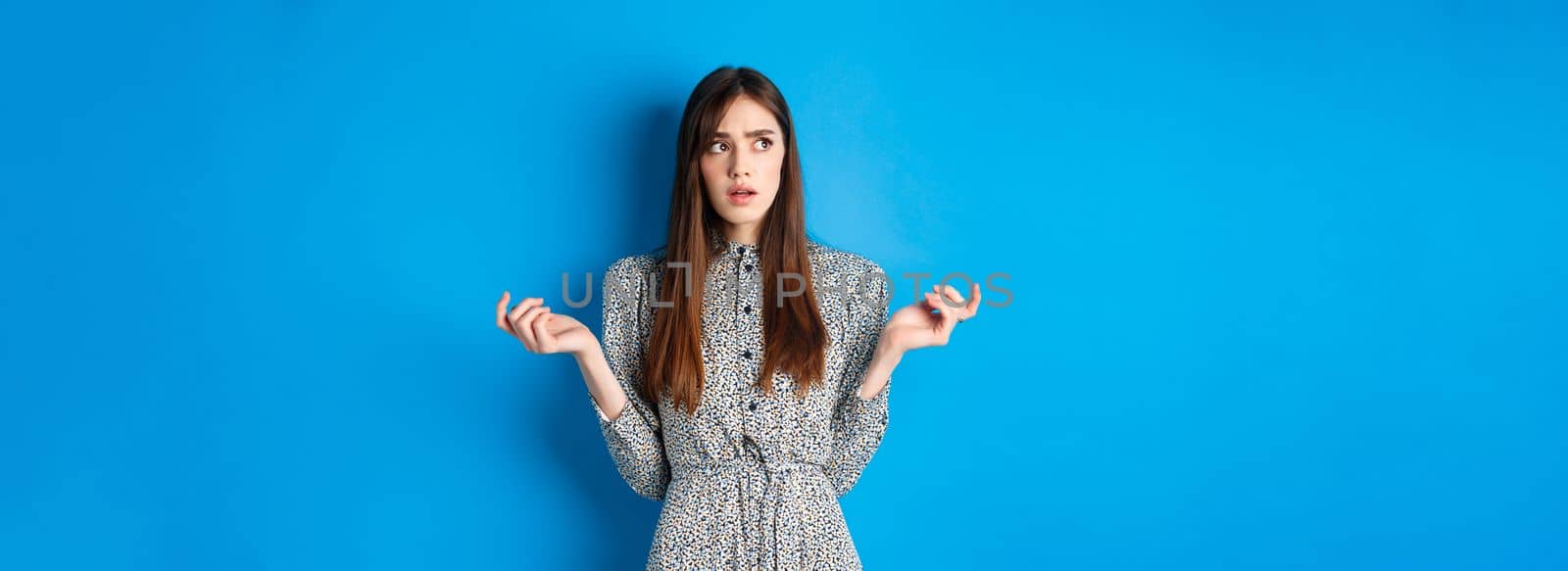 Confused cute girl cant understand something, look away pensive and counting on fingers, standing in dress against blue background by Benzoix