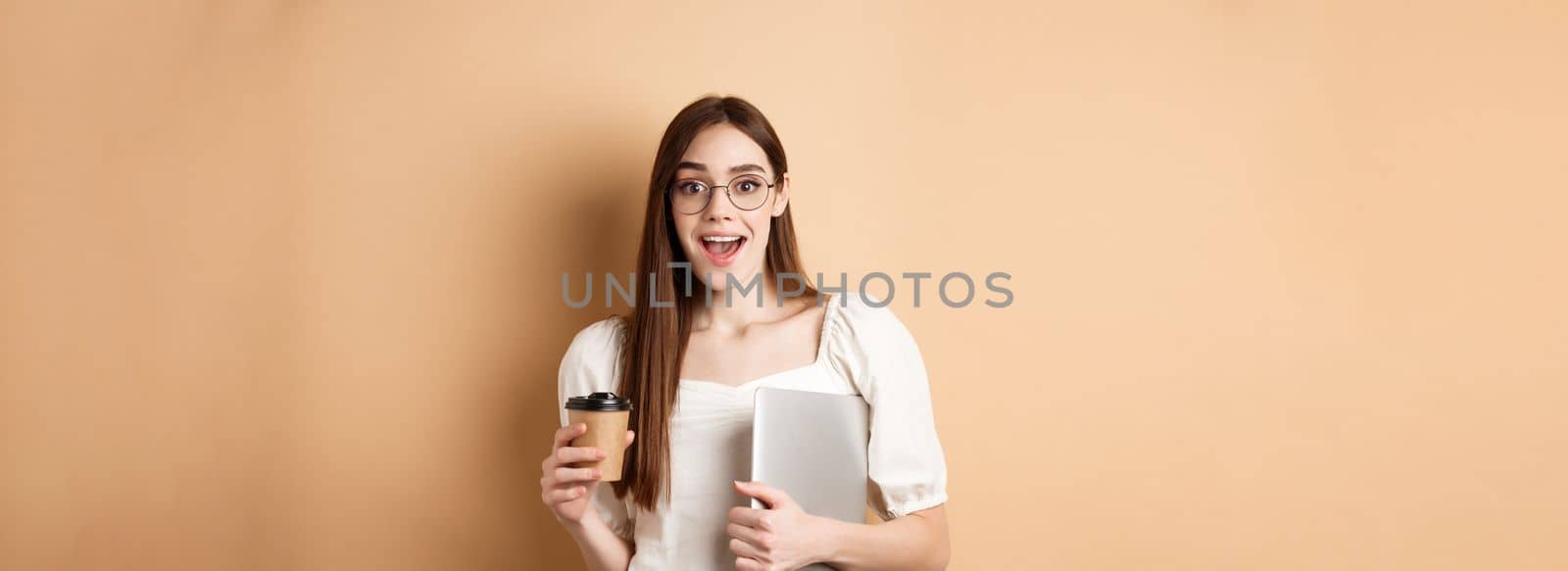 Excited woman in glasses drinking coffee from takeout cup, holding laptop and smiling happy, standing on beige background by Benzoix