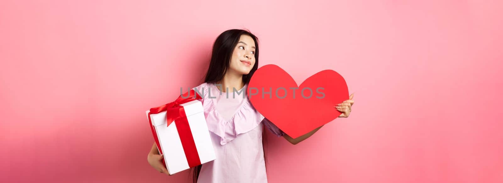 Valentines day concept. Romantic girl holding lover gifts, big red heart card and box with present, looking aside with dreamy look, falling in love, smiling tenderly, pink background.