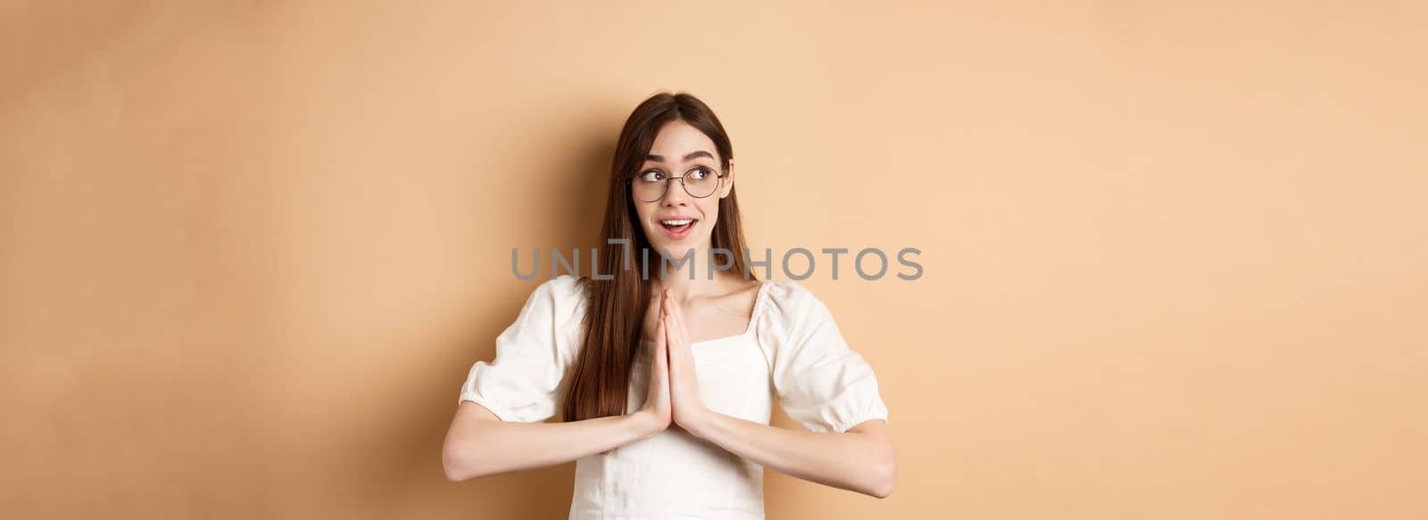 Hopeful girl holding hands in pray, begging for dream come true, looking aside excited, standing on beige background. Copy space