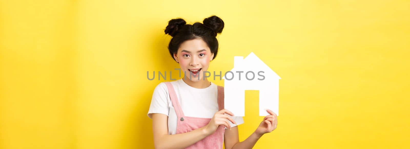 Real estate and family concept. Cute asian woman with bright makeup and stylish hairbuns, showing paper house cutout, smiling determined, yellow background.