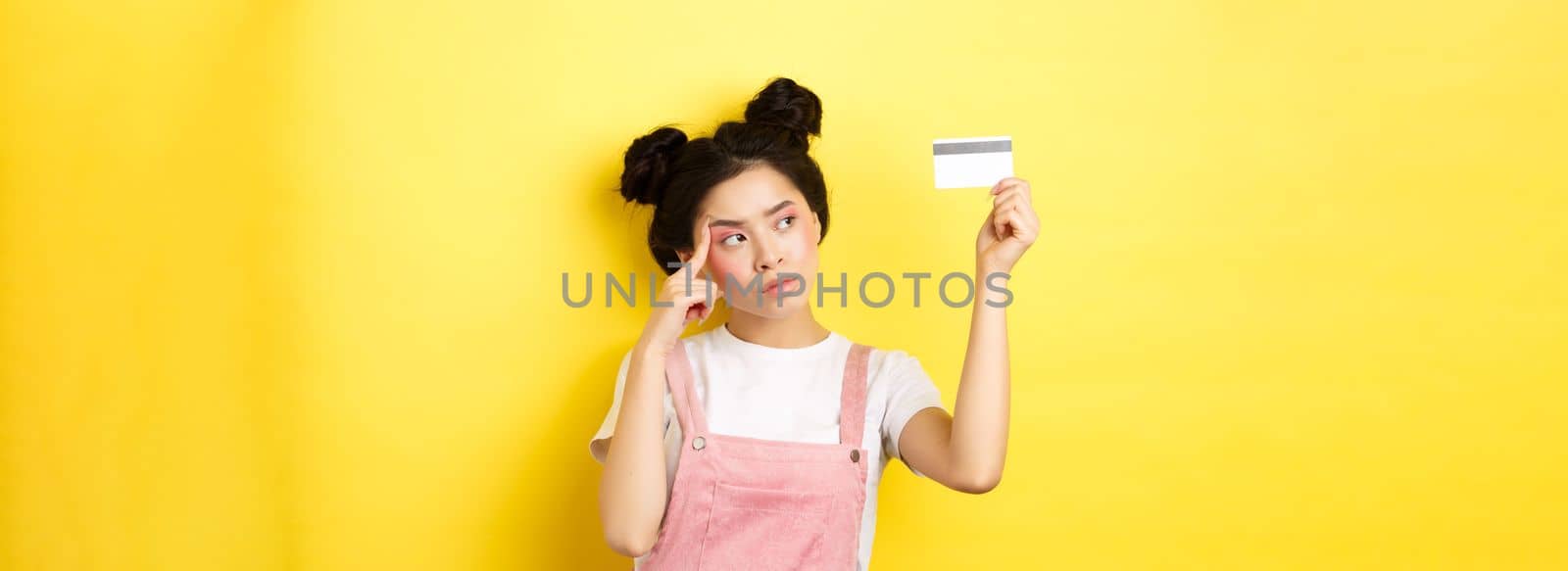 Shopping. Thoughtful trendy girl with summer makeup, looking pensive at plastic credit card, standing on yellow background.