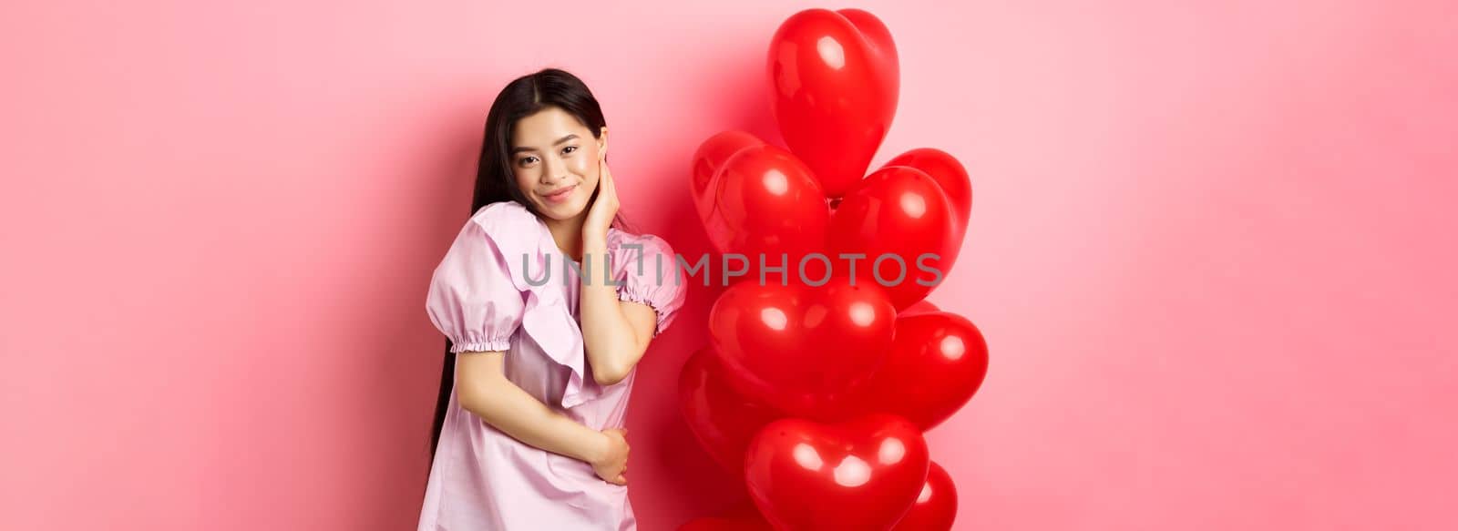Beautiful asian girl in dress smiling coquettish, flirting on valentines day, looking sensual at camera, posing near valentines heart balloons, pink background by Benzoix