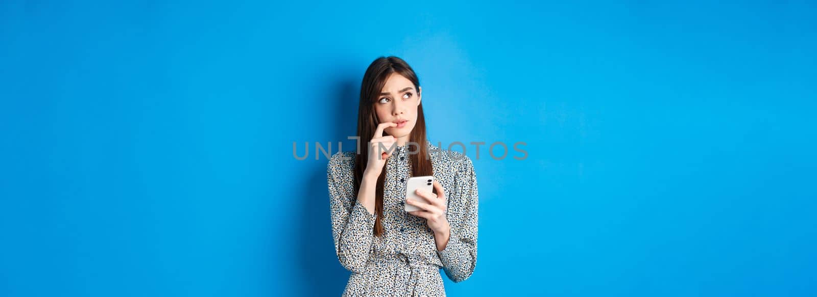 Pensive cute girl thinking how to answer on message, looking aside thoughtful and holding smartphone, standing in dress on blue background by Benzoix