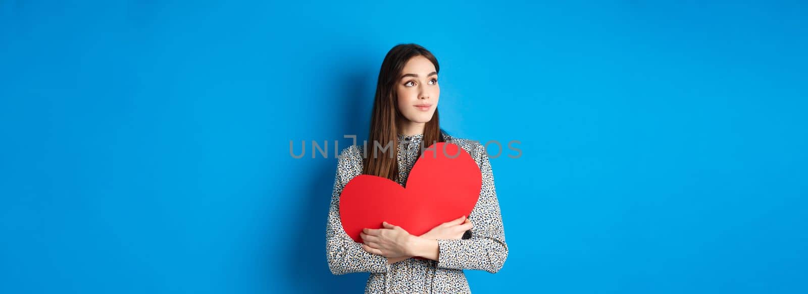 Valentines day. Romantic girl looking dreamy at upper left corner and smiling, holding big red heart cutout, standing on blue background.