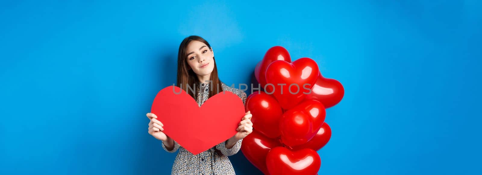 Valentines day. Romantic girl in dress showing big red heart cutout, dreaming of love, standing near holiday balloons on blue background.