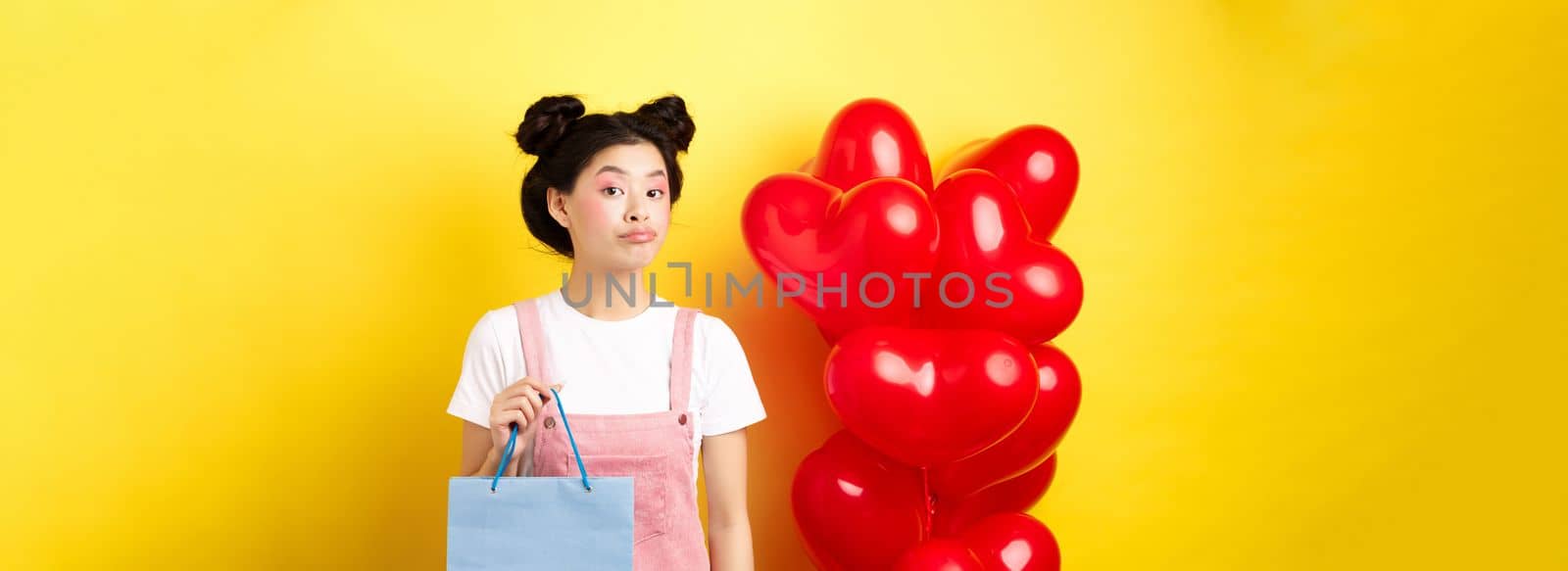 Happy Valentines day. Stylish asian single girl buying herself gift, holding shopping bag and looking unbothered at camera, standing over yellow background by Benzoix