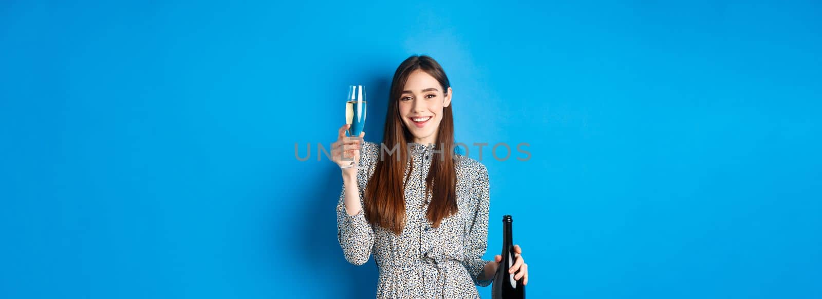 Celebration. Happy young woman in dress raising glass of champagne, holding bottle and smiling, having fun at party, standing on blue background by Benzoix