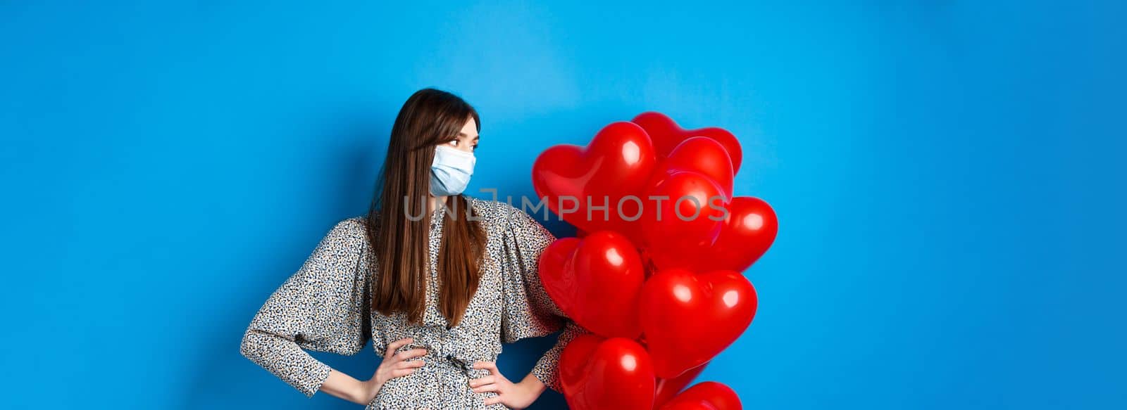 Covid-19 and Valentines day. Young bothered girl in medical mask and dress, looking at heart balloons, waiting on date, standing blue background.