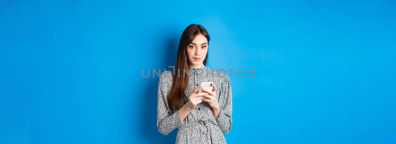 Stylish modern woman in dress, chatting on smartphone and smiling at camera, standing on blue background by Benzoix