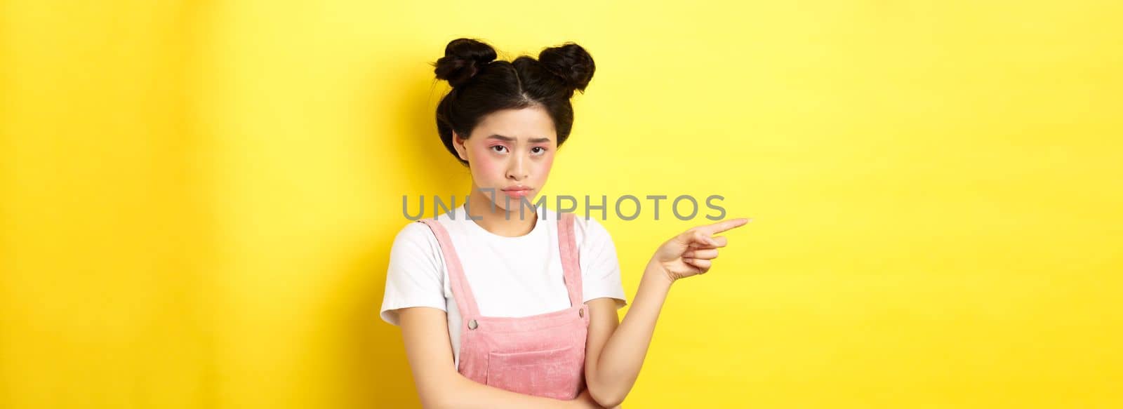 Sad and gloomy asian girl with glamour makeup, frowning unfair and pointing left at logo, looking disappointed, standing on yellow background.